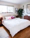 Cozy bedroom with a large bed, colorful pillows, wooden dresser, framed art, and a potted plant by the window.