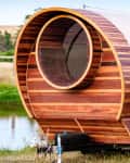 Wooden barrel-shaped tiny house on wheels with a large circular window, set in a grassy landscape near a pond.