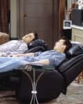 Two people reclining on black leather chairs in a living room with a brown sofa and glass side table.