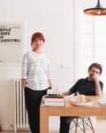 Man sitting at a wooden desk with books and camera, woman standing beside, modern art on wall, pendant light above.