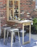 Outdoor bar setup with wooden shelf, metal stools, plants, and drinks on a blue patterned rug against a brick wall.