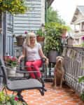 Woman in white shirt and red pants sitting on a balcony with a dog, surrounded by potted plants and flowers.