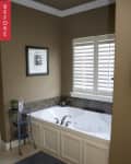 Bathroom with a white corner bathtub, beige walls, metal shelf, and shuttered window.
