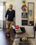 Man in a navy shirt and khaki pants stands in a modern living room with eclectic decor, including a "Cool Beans" pillow.