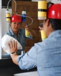 Person cleaning a mirror wearing a red helmet with attached cleaning bottles, using a cloth and spray.