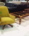 Furniture showroom with a yellow swivel chair, glass table, leafy plant, and wooden coffee table.