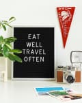 Letter board with "Eat Well Travel Often," a potted plant, vintage camera, and red pennant on a white surface.