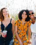 Three women smiling outdoors, one holding a drink, standing in front of tall grass.