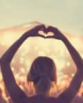 Concert crowd with person making a heart shape with hands against a brightly lit stage.