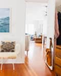 White room with ocean artwork, a chair with a patterned cushion, and a small table with books and a lamp. Open door to living area.