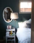Small bedroom with teal walls, white bedding, oval mirror, and bedside table with lamp and photo frame.