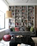 Living room with a gray sofa, colorful cushions, large bookshelf, brick wall, and a black coffee table.