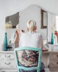 Person with short hair applying makeup at a vintage vanity with a tri-fold mirror and decorative chair.