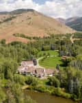 Large estate surrounded by lush trees and a river, set against rolling hills and mountains under a partly cloudy sky.