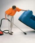Person in orange shirt vacuuming under a blue sofa with a red vacuum cleaner.
