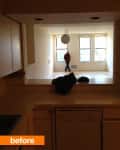 View from kitchen to empty living room with large windows and a hanging light fixture, person walking in the distance.