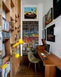 Home office with wooden desk, computer, bookshelves, yellow lamp, and artwork on walls.