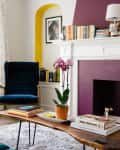 Living room with purple accent wall, bookshelves, blue chair, wooden coffee table, orchid plant, and a white fireplace.