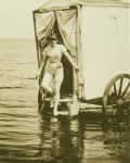 Woman stepping out of a vintage bathing machine into the sea, wearing a swimsuit.