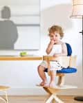 Toddler in a wooden high chair at a table with grapes, next to a floor lamp and abstract wall art.