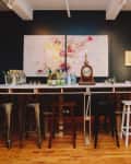 Bar setup with stools, abstract art, wine bottles, and a vintage clock on a wooden counter.