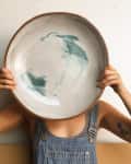 Person in overalls holding a large ceramic plate with green abstract design in front of their face.