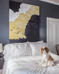 Dog sitting on a white bed in a dark-walled bedroom with abstract art, a side table, and a chandelier.