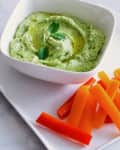 Creamy green dip garnished with a mint leaf, served with orange and red bell pepper sticks on a white plate.