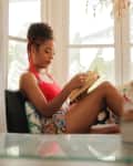 Woman in red top and floral shorts reading a book by a window, with a stack of books and a pen holder nearby.