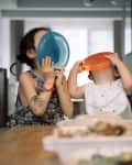 Two children playfully holding colorful plates over their faces at a dining table with food.
