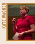 Person in a red dress speaking on stage, holding a paper and microphone, with "Kate Wagner 2020" text on the side.