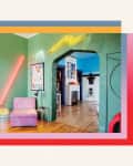 Eclectic living room with green walls, neon lights, pink chair, and art posters, leading to a bedroom and a blue-walled room.