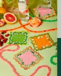 Colorful embroidered coasters with fruit and drinks on a green surface, alongside a vibrant abstract puzzle and modern chandelier.