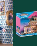 A Playmobil figure in a blue outfit with a pink hat and handbag stands in a patterned room, next to a dollhouse box.