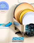 Dish rack with colorful plates, glasses, and utensils drying next to a sink, with a sponge and dish soap nearby.