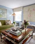 Cozy living room featuring a green sofa with patterned pillows, a wooden coffee table stacked with books, and floral decor.