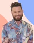 Man with a beard wearing a colorful abstract-patterned jacket, standing against a pastel geometric background.