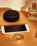 Smart speaker, smartphone, perfume bottle, and jewelry on a wooden table with a decorative pillow.