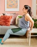 Woman doing tricep dips on a chair in a colorful living room with a gray sofa and decorative pillows.