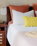 Bed with pink and white pillows, yellow striped cushion, wooden nightstand, lamp, plant, and folded linens.