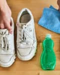 White sneakers on wooden floor with blue cloth, green detergent, sponge, and toothbrush for cleaning.