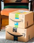 Two Amazon Prime packages stacked on a rug in a living room with a chair and plant nearby.
