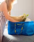 Woman placing pillows and a plant into a blue IKEA bag on a bed with colorful cushions.