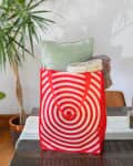 Red shopping bag with spiral design on table, filled with a green pillow and a colorful blanket, surrounded by plants.