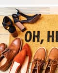 Doormat with "OH, HELLO" surrounded by various shoes, including heels, sneakers, and loafers.