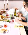 Friends enjoying a meal with noodles, dumplings, and wine at a dining table with pink tablecloth and white flowers.