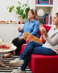 Four women enjoying snacks and drinks in a cozy living room with a small dog on a red chair.