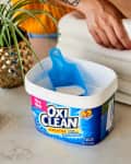 OxiClean container with scoop on counter, next to a jar of clothespins, a plant, and folded white towels.