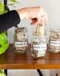 Hand placing cash into a jar labeled "House Fund" among jars for concert, travel, and class savings on a wooden shelf.
