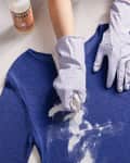 Person wearing gloves cleaning a blue sweater with paint thinner and dish soap on a white surface.